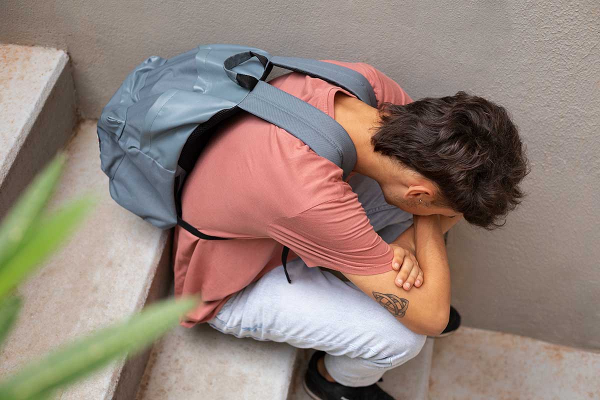 A young man sits on stairs and crouches over, looking at his arms