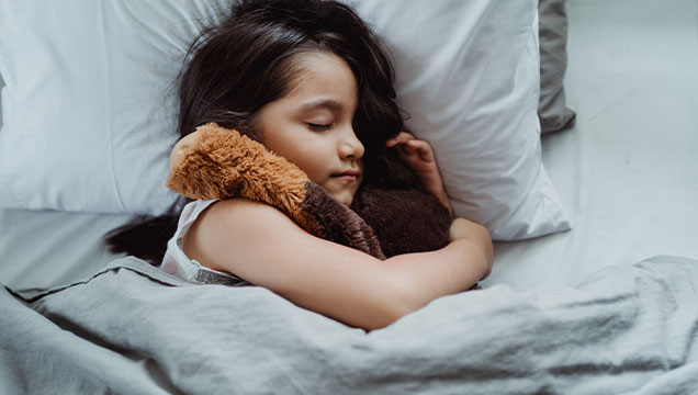 Child resting their head on a pillow with their eyes closed on a bed with the doona pulled up to under their arm, embracing a teddy bear.