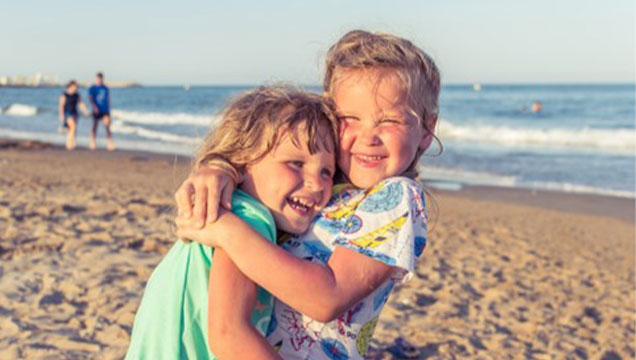 Young siblings at the beach embracing one another and smiling.