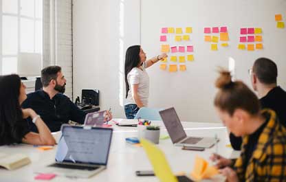 A person in a brightly lit room pointing at a wall covered in sticky notes with other people sitting at a desk with laptops looking in the same direction