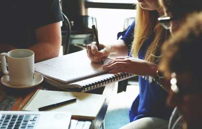 Group of people sitting at a wooden table with notepads, laptops and coffee mug and saucer