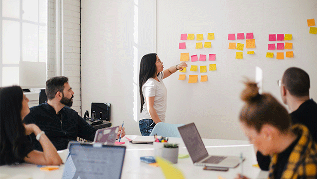 person in a brightly lit room pointing at a wall covered in sticky notes with other people sitting at a desk with laptops looking in the same direction