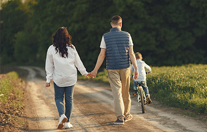 Mother and Father walk along holding hands while their son rides his bike in the distance.