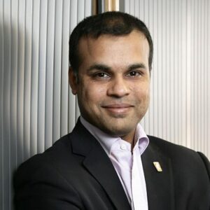 Dr Raja Mukherjee, UK-based psychiatrist, short black hair, pale collared shirt and black blazer, smiling warmly at the camera against a backdrop of white vertical blinds.