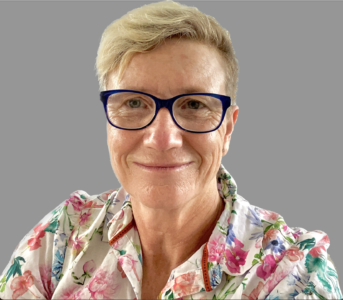 Vanessa Spiller, clinical psychologist, short blonde hair, floral blouse and navy-rimmed glasses, smiling warmly against a grey background.